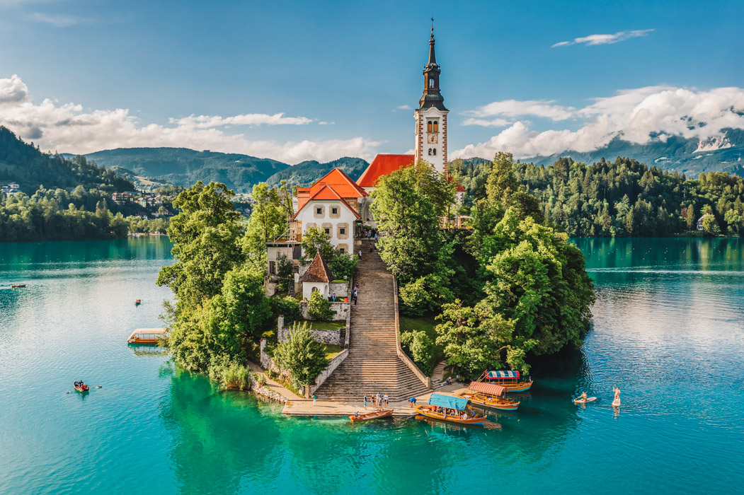 Bled, Slovenia - Aerial view of beautiful Pilgrimage Church of the Assumption of Maria on a small island at Lake Bled (Blejsko Jezero) and lots of Pletna boats on the lake at summer time with blue sky